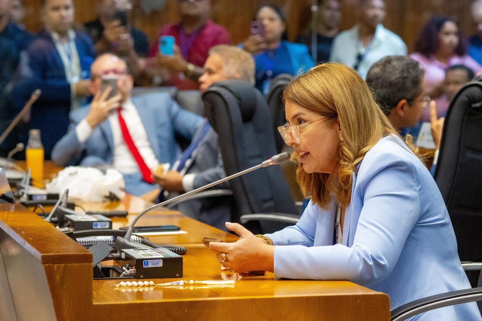 Iracema Vale, falando ao microfone e segurando um celular, durante sua reeleição na Assembleia Legislativa do Maranhão, enquanto Othelino Neto aparece ao fundo falando ao celular.