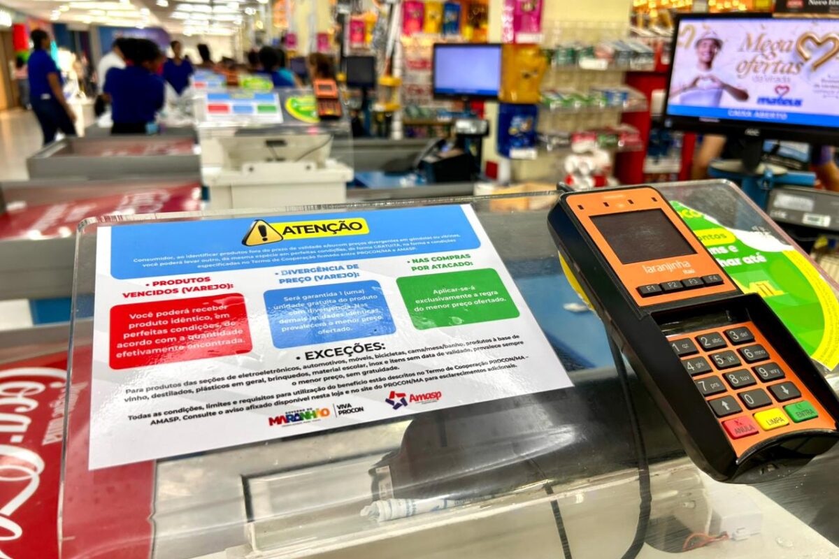 Aviso informativo do Procon-MA sobre direitos do consumidor em caso de divergência de preços, produto vencido e compra por atacado afixado no caixa de um supermercado, ao lado de uma máquina de cartão.
