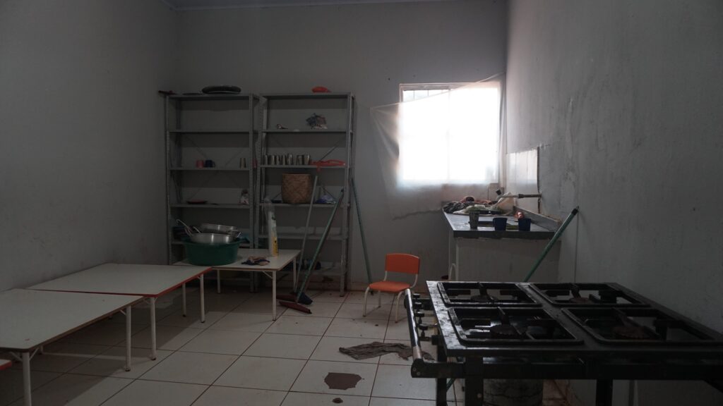 Foto em plano aberto de um ambiente interno usado como cozinha. O chão de cerâmica branca está sujo, com manchas escuras e um pano de chão velho jogado no centro. À direita, um botijão de gás está no chão conectado a uma mangueira. Ao fundo, há uma pia de cimento com louças e produtos misturados. Vassouras estão encostadas nas paredes e uma pequena cadeira infantil laranja está isolada no meio da sala.