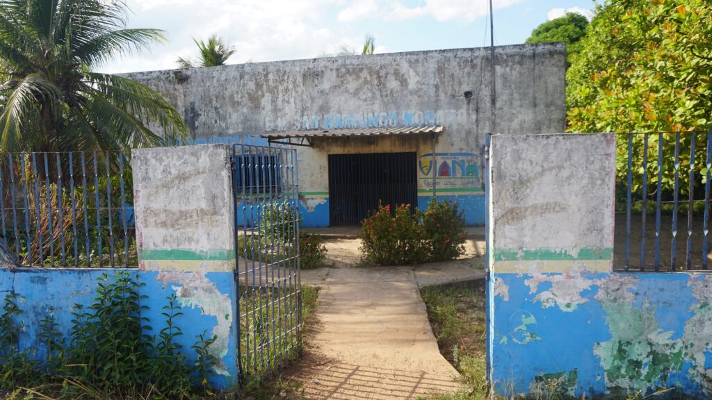 Foto colorida horizontal mostrando a frente da Escola Municipal São Raimundo Nonato. O prédio é térreo, pintado de branco com uma faixa azul na base, mas a tinta está muito desgastada e descascando, dando um aspecto envelhecido. No alto da parede, lê-se o nome da escola em letras azuis desbotadas. Em primeiro plano, um portão de grades azuis está aberto, revelando um caminho de cimento que leva à entrada escura da sala de aula, protegida por uma grade preta. Mato cresce ao redor do muro e dentro do pátio de entrada.