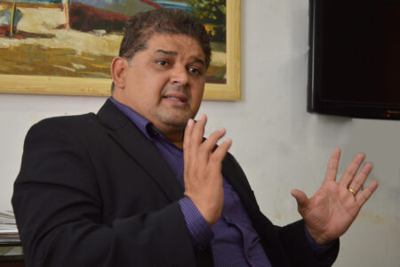 Homem de terno preto e camisa roxa gesticula com as duas mãos enquanto fala. Ao fundo, quadro com pintura e TV na parede.