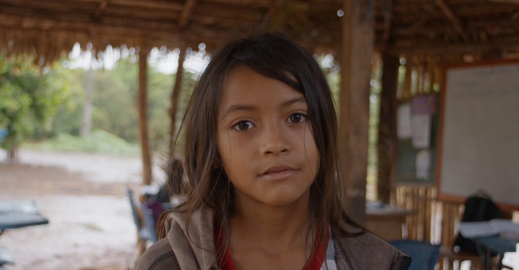 Retrato de uma menina indígena com cerca de nove anos, cabelos longos e soltos, olhando para a câmera. Ao fundo, vê-se o interior de uma sala de aula improvisada, com estrutura de madeira e palha, mesas simples e o ambiente aberto para a área externa da aldeia.