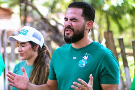 Homem de barba gesticula com as mãos abertas, vestindo camiseta verde com adesivos de campanha do número 44. Ao fundo, mulher de boné branco e camiseta verde.