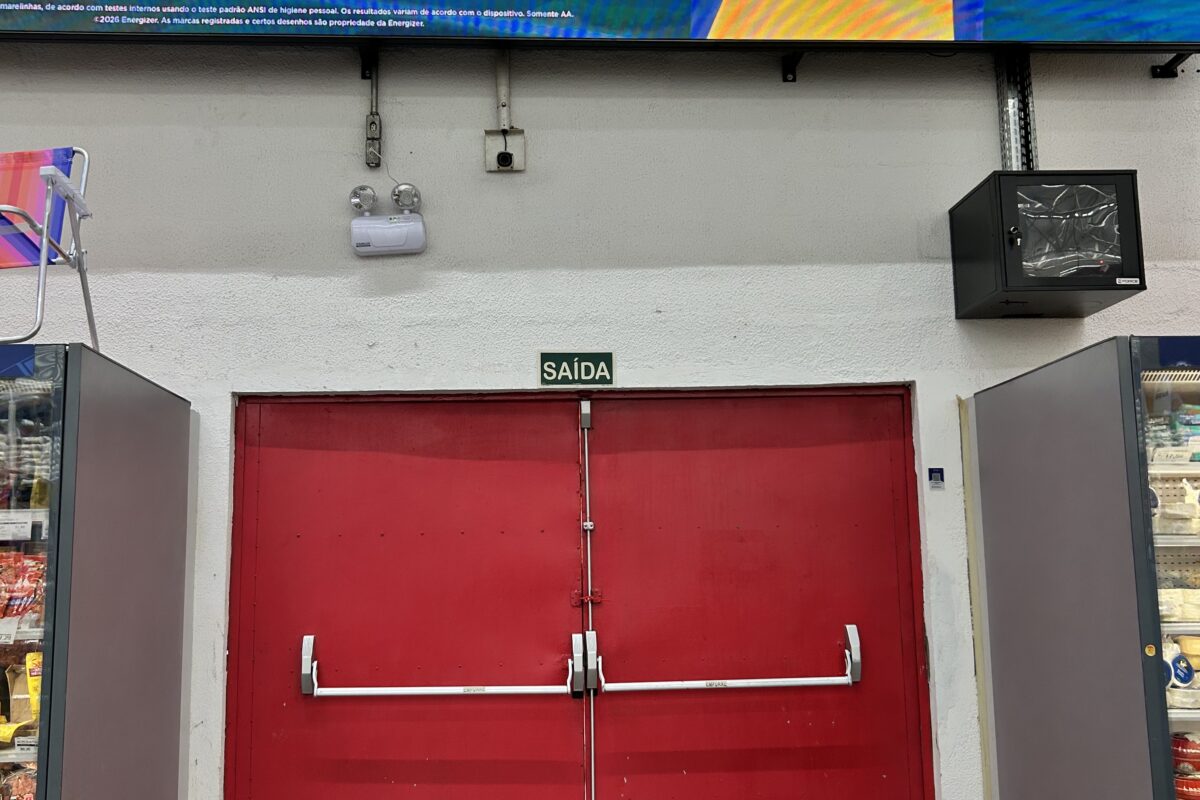 Porta dupla vermelha de saída de emergência na unidade do Grupo Mateus da Curva do 90, em São Luís, entre dois balcões refrigerados, com placa de "Saída" acima da porta e um painel eletrônico de publicidade na parte superior da imagem.