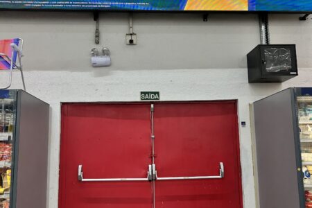 Porta dupla vermelha de saída de emergência na unidade do Grupo Mateus da Curva do 90, em São Luís, entre dois balcões refrigerados, com placa de "Saída" acima da porta e um painel eletrônico de publicidade na parte superior da imagem.