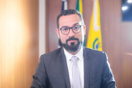 Homem de barba e óculos, em terno escuro e gravata clara, olhando para a câmera. Atrás dele, bandeiras hasteadas.