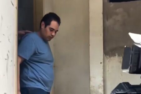 Homem de camiseta azul e cabelo escuro, cabeça baixa, parado em uma porta com uma mão pousada sobre seu ombro. À direita, câmera de vídeo apontada para ele.