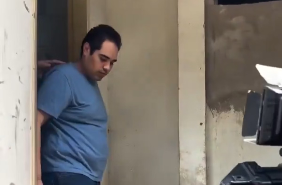 Homem de camiseta azul e cabelo escuro, cabeça baixa, parado em uma porta com uma mão pousada sobre seu ombro. À direita, câmera de vídeo apontada para ele.
